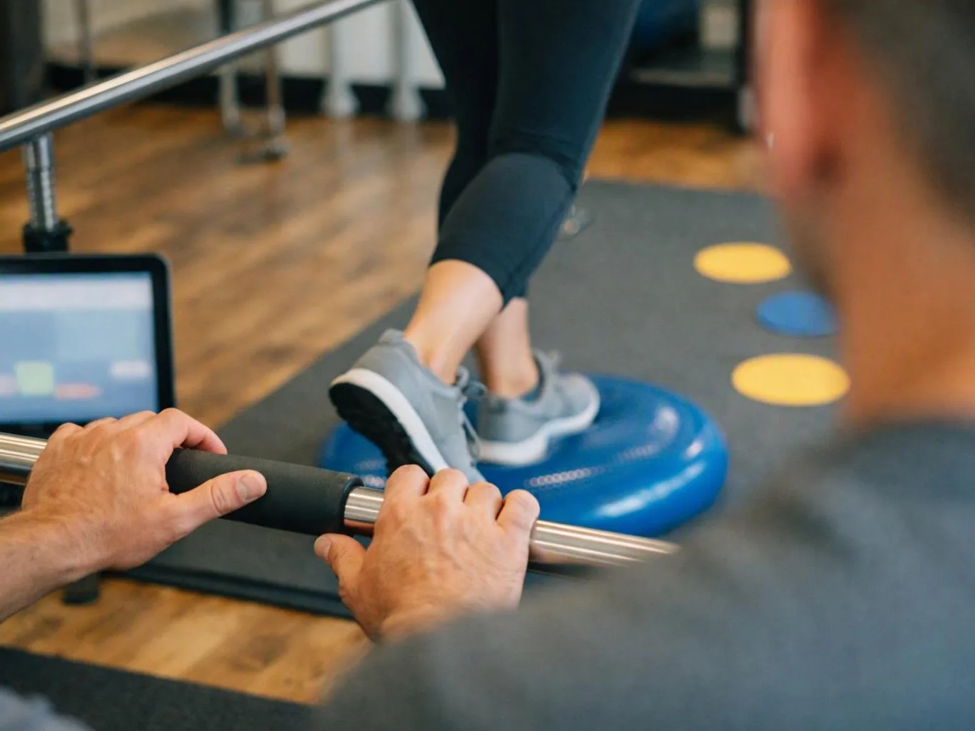 A person balances on a blue wobble disc while another grips a metal rail in a gym therapy room