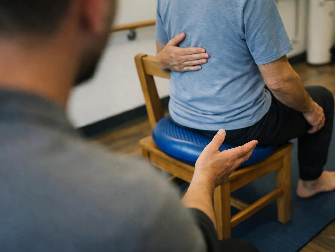 A therapist guides a seated man with hand placements on his back and shoulder in a clinic room