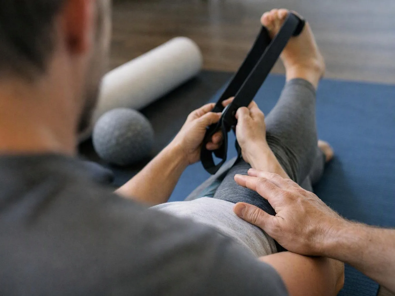 A therapist stretches a person’s raised leg with a strap on a blue exercise mat